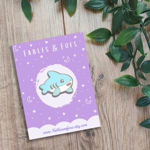 May include: A light blue shark enamel pin with white detailing and a smiling face. The pin is displayed on a purple card with the text "FABLES & FOES" and a website address. The background is a wooden surface with green leaves.