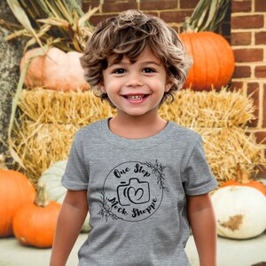 May include: A child wearing a grey t-shirt with a black circular logo reading "One Step Mock Shoppe". The background features pumpkins, straw bales, and a brick wall, creating an autumnal atmosphere.
