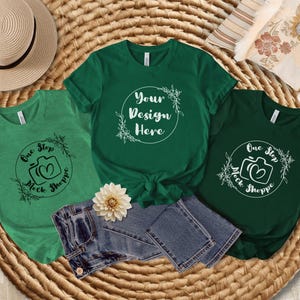 May include: Three green t-shirts with different designs. The center shirt says "Your Design Here" with a floral design. The left shirt says "One Step Mock Shoppe" with a camera graphic. The right shirt says "One Step Mock Shoppe" with a camera graphic.