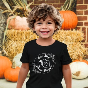 May include: A young child wearing a black t-shirt with a white graphic design that says "One Stop Mock Shoppe" and a camera icon. The child is standing in front of a brick wall with pumpkins in the foreground.