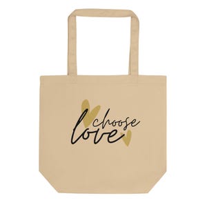 May include: Beige tote bag with a rectangular shape and two handles. The bag features the words "choose love" in black script with gold accents. The bag is made of a canvas-like material.