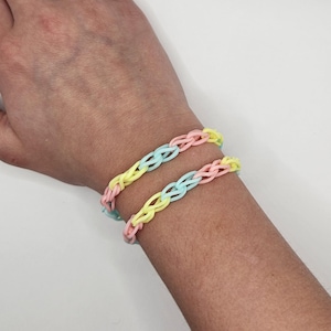 May include: Two pastel-colored rubber band bracelets are shown on a person's wrist. The bracelets are made of interlocking rubber bands in shades of pink, yellow, and blue. The bracelets are a fun and colorful accessory.