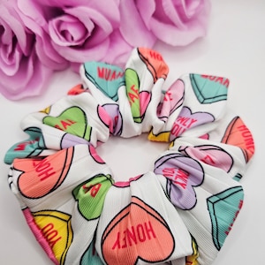 May include: White scrunchie with a colorful candy heart pattern. The hearts have various phrases on them, including "Honey", "You're My", "Bae", "Only You", and "Wifey".