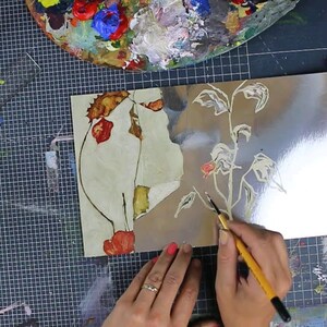 Puede incluir: Una persona está pintando un diseño floral en una superficie plateada usando un pincel pequeño. El diseño es de una planta con hojas y flores. La persona lleva un anillo en su dedo.