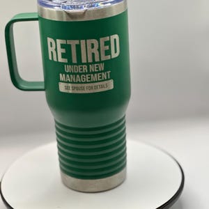 May include: Green stainless steel travel mug with a lid and handle. The mug has the text "Retired Under New Management See Spouse for Details" on the side.