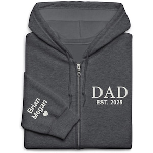 May include: A dark charcoal gray zip-up hoodie featuring white text on the chest that reads DAD EST. 2025. The sleeve is embroidered with the names Brian and Megan followed by a small white heart symbol. The garment has a hood and drawstrings.