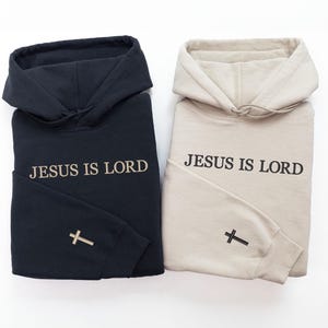 Puede incluir: Se muestran dos sudaderas con capucha, una negra y otra beige. El texto "JESUS IS LORD" est&aacute; impreso en la parte delantera de cada sudadera. Una cruz est&aacute; bordada en la manga izquierda de cada sudadera. Las sudaderas est&aacute;n dobladas.