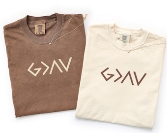 May include: Two short-sleeved crew neck t-shirts. One is brown with cream-colored text reading G>AV, and the other is cream with brown text reading G>AV. Both shirts feature a Comfort Colors brand label at the inner neckline and a thin gold chain.