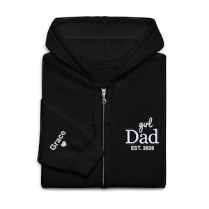 May include: A black zip-up hoodie featuring white embroidered text on the chest that reads girl Dad EST. 2026. The left sleeve cuff displays the name Grace followed by a small white heart icon. The garment includes a hood and a full-length zipper.