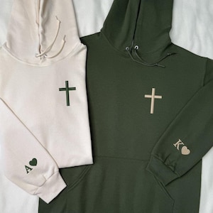May include: Two hooded sweatshirts, one cream and one dark green. Each has a cross embroidered on the chest. The cream hoodie has an embroidered heart and the letter "A" on the sleeve. The green hoodie has a heart and the letter "K" on the sleeve.