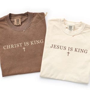 K&ouml;nnte beinhalten: Zwei kurz&auml;rmelige T-Shirts mit Rundhalsausschnitt. Das braune Shirt zeigt den Schriftzug "CHRIST IS KING" mit einem Kreuz in hellem Beige. Das cremefarbene Shirt zeigt "JESUS IS KING" mit einem Kreuz in Dunkelbraun. Beide haben Comfort Colors Etiketten.
