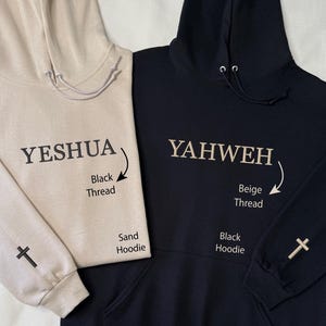 May include: Two hooded sweatshirts, one sand-colored and one black. The sand hoodie has "YESHUA" embroidered in black thread, with a cross on the sleeve. The black hoodie features "YAHWEH" in beige thread, also with a cross on the sleeve.