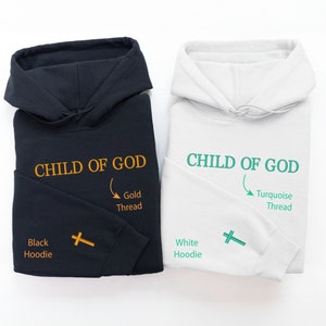 Op de afbeelding: Twee hoodies, een zwarte en een witte, met de tekst "CHILD OF GOD" geborduurd op de voorkant. De zwarte hoodie heeft borduurwerk met gouden draad en de witte hoodie heeft borduurwerk met turquoise draad. Beide hoodies hebben een kruis geborduurd op de mouw.