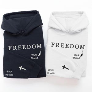 May include: Two folded hoodies, one black and one white. The word "FREEDOM" is printed in large letters on the front of each hoodie. A small cross is embroidered on the left sleeve. The black hoodie has white thread, and the white hoodie has black thread.