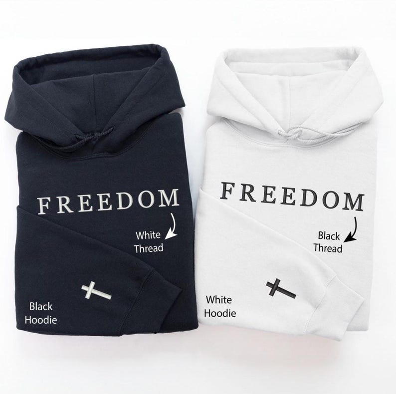 May include: Two folded hoodies, one black and one white. The word "FREEDOM" is printed in large letters on the front of each hoodie. A small cross is embroidered on the left sleeve. The black hoodie has white thread, and the white hoodie has black thread.