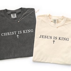 K&ouml;nnte beinhalten: Ein anthrazitfarbenes T-Shirt mit dem wei&szlig;en Text CHRIST IS KING und einem Kreuz sowie ein cremefarbenes T-Shirt mit dem schwarzen Text JESUS IS KING und einem Kreuz. Beide T-Shirts haben einen Rundhalsausschnitt und ein Comfort Colors Etikett.