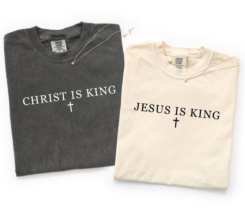 K&ouml;nnte beinhalten: Ein anthrazitfarbenes T-Shirt mit dem wei&szlig;en Text CHRIST IS KING und einem Kreuz sowie ein cremefarbenes T-Shirt mit dem schwarzen Text JESUS IS KING und einem Kreuz. Beide T-Shirts haben einen Rundhalsausschnitt und ein Comfort Colors Etikett.