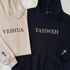 May include: Two hooded sweatshirts, one beige and one black. The beige sweatshirt has the word "YESHUA" embroidered on the front and a cross on the sleeve. The black sweatshirt has "YAHWEH" embroidered on the front and a cross on the sleeve. Both have drawstrings.