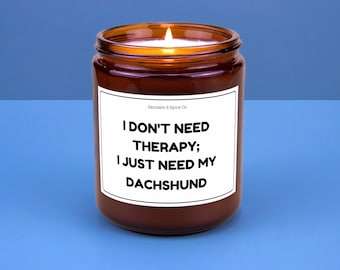 May include: A lit brown glass jar candle with a white label. The label reads "I DON'T NEED THERAPY; I JUST NEED MY DACHSHUND" in black text. The candle is on a blue surface.