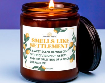 May include: A 9oz soy wax candle in an amber glass jar with a black lid. The label features lemon illustrations and the text: SMELLS LIKE SETTLEMENT, A SWEET SCENT REMINISCENT OF THE DIVISION OF ASSETS AND THE SPLITTING OF A ONCE SHARED LIFE.