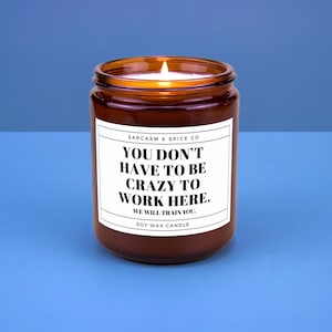 May include: A brown glass jar candle with a white label. The label reads "YOU DON'T HAVE TO BE CRAZY TO WORK HERE. WE WILL TRAIN YOU." The candle is a soy wax candle.