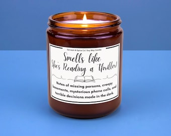 May include: A brown glass jar candle with a lit wick. The label reads "Smells like She's Reading a Thriller!" with text about missing persons and creepy basements. The candle sits on a blue surface.