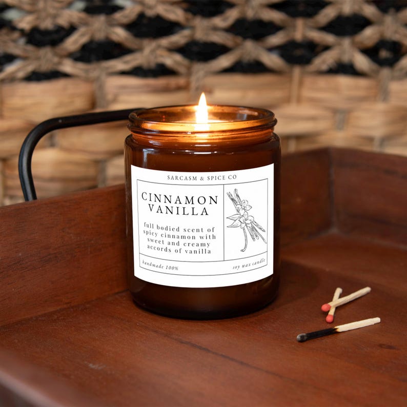K&ouml;nnte beinhalten: Ein braunes Glasgef&auml;&szlig; mit einer Kerze und einem wei&szlig;en Etikett, auf dem "SARCASM & SPICE CO" und "CINNAMON VANILLA" steht. Das Etikett beschreibt den Duft auch als "vollmundiger Duft von w&uuml;rziger Zimt mit s&uuml;&szlig;en und cremigen Vanille-Noten". Die Kerze ist angez&uuml;ndet und es liegen drei Streichh&ouml;lzer auf einer Holzoberfl&auml;che.