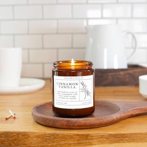 K&ouml;nnte beinhalten: Ein braunes Glasgef&auml;&szlig; mit einer Kerze und einem wei&szlig;en Etikett, auf dem "SARCASM & SPICE CO" und "CINNAMON VANILLA" steht. Das Etikett enth&auml;lt auch eine Beschreibung des Duftes: "Full bodied scent of spicy cinnamon with sweet and creamy accords of vanilla". Die Kerze ist angez&uuml;ndet und die Flamme ist sichtbar.
