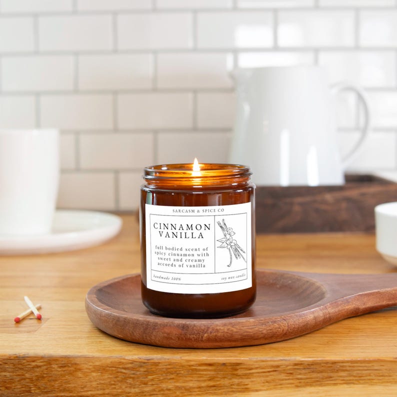K&ouml;nnte beinhalten: Ein braunes Glasgef&auml;&szlig; mit einer Kerze und einem wei&szlig;en Etikett, auf dem "SARCASM & SPICE CO" und "CINNAMON VANILLA" steht. Das Etikett enth&auml;lt auch eine Beschreibung des Duftes: "Full bodied scent of spicy cinnamon with sweet and creamy accords of vanilla". Die Kerze ist angez&uuml;ndet und die Flamme ist sichtbar.
