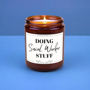 May include: A brown glass jar candle with a white label. The label reads "DOING Social Worker STUFF" in black script and block letters. The candle has a lit wick and is set against a blue background.