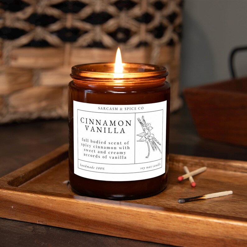K&ouml;nnte beinhalten: Ein braunes Glasgef&auml;&szlig; mit einer Kerze und einem wei&szlig;en Etikett, auf dem "SARCASM & SPICE CO" und "CINNAMON VANILLA" steht. Das Etikett enth&auml;lt auch eine Beschreibung des Duftes: "full bodied scent of spicy cinnamon with sweet and creamy accords of vanilla". Die Kerze ist angez&uuml;ndet und die Flamme ist sichtbar.