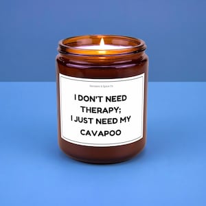May include: A lit brown glass jar candle with the text "I DON'T NEED THERAPY; I JUST NEED MY CAVAPOO" on a white label. The candle is on a blue surface. The brand name "Sarcasm & Spice Co" is at the top of the label.