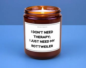 May include: A lit brown glass jar candle with a white label. The label reads "I DON'T NEED THERAPY; I JUST NEED MY ROTTWEILER" in black text. The candle is on a blue surface.