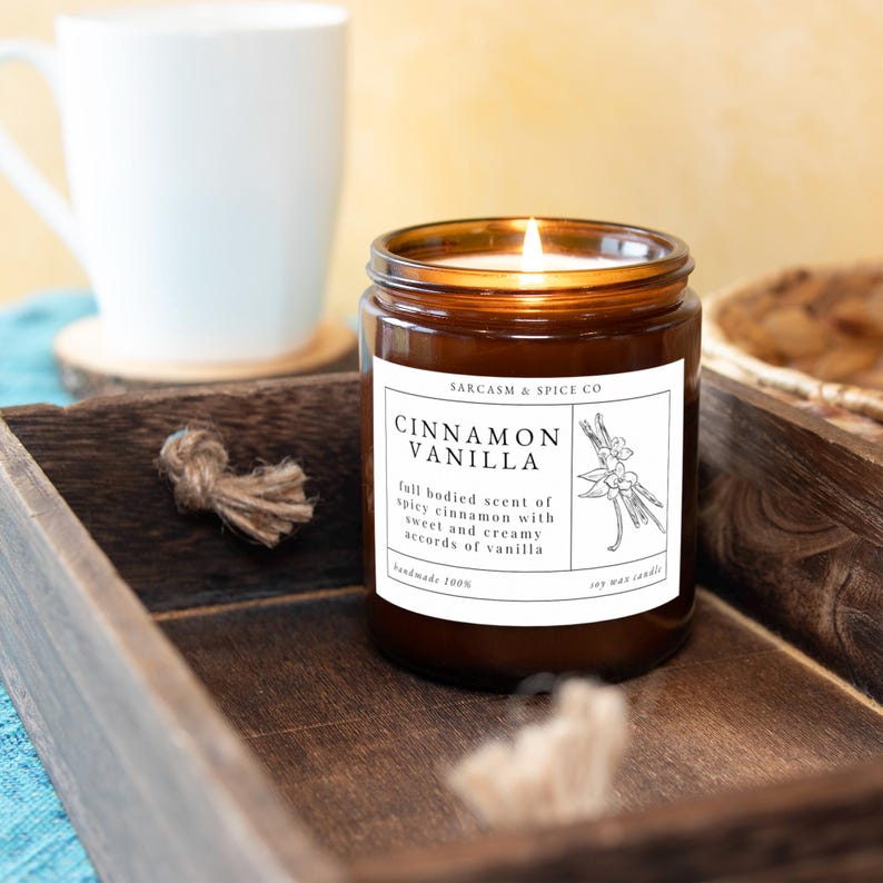 K&ouml;nnte beinhalten: Eine braune Glaskerze mit einem wei&szlig;en Etikett, auf dem "SARCASM & SPICE CO" und "CINNAMON VANILLA" steht. Das Etikett enth&auml;lt auch eine Beschreibung des Duftes: "vollmundiger Duft von w&uuml;rziger Zimt mit s&uuml;&szlig;en und cremigen Vanille-Noten". Die Kerze ist angez&uuml;ndet und die Flamme ist sichtbar.
