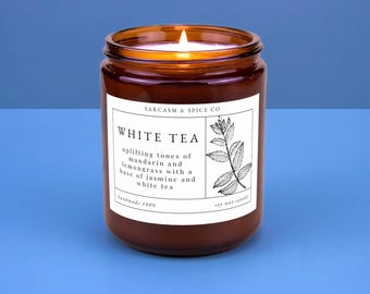 May include: A brown glass candle jar with a white label that reads "SARCASM & SPICE CO" and "WHITE TEA". The label also describes the scent as "uplifting tones of mandarin and lemongrass with a base of jasmine and white tea". The candle is lit and the wax is melting.