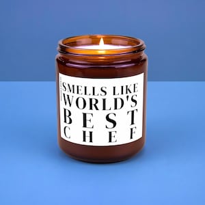 May include: A lit brown glass jar candle with a white label. The label reads "Smells Like World's Best Chef" in bold black letters. The candle sits on a blue surface.