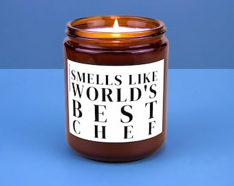 May include: A lit brown glass jar candle with a white label. The label reads "Smells Like World's Best Chef" in bold black letters. The candle sits on a blue surface.