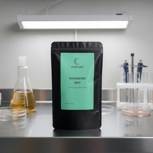 May include: A black pouch with a teal label displaying the text "Culture Clarity Potato Dextrose Broth" sits on a stainless steel surface. Laboratory equipment, including beakers, petri dishes, and test tubes, are in the background.