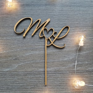 Könnte beinhalten: Holz-Kuchentopper mit den Buchstaben "M & L" in einer kursiven Schriftart mit einem Herzen zwischen den Buchstaben. Der Topper ist auf einem Holzstab montiert.