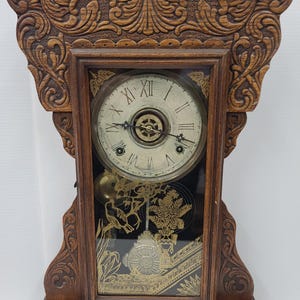 May include: A vintage wooden clock with an ornate, carved design. The clock face has Roman numerals and decorative hands. The clock's glass panel displays a gold-colored illustration of birds and foliage. The clock is a dark brown color.