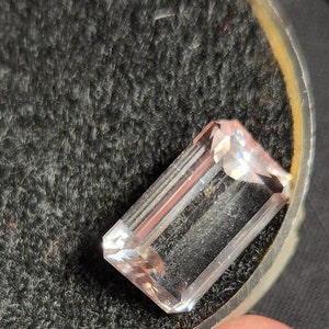 May include: A clear, faceted, rectangular gemstone with a slight pink hue. The gemstone is sitting in a black velvet box.