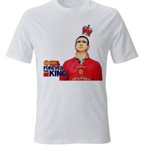 Puede incluir: Camiseta blanca con una imagen roja y blanca de un jugador de fútbol que lleva una camiseta roja con el texto "UMBRO" y "SHARP" en ella. El jugador lleva una corona en la cabeza. El texto "ERIC CANTONA FOREVER THE KING" está impreso en la camiseta.