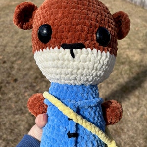 May include: A crocheted plush toy in the shape of an otter wearing a blue jacket with a yellow strap. The otter has brown fur, white fur around its mouth, and black eyes.