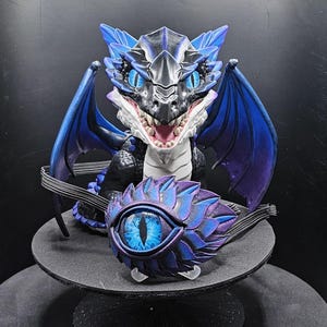 May include: A detailed figurine of a dragon with blue and purple wings, scales, and eyes. The dragon is perched on a round, gray platform. A separate, eye-shaped object with a blue iris is also present.