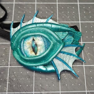 May include: A teal and silver eye mask with a detailed, reptilian design. The mask features a large, realistic eye with blue and gold tones, surrounded by textured teal scales and fin-like accents. A black elastic band is attached.