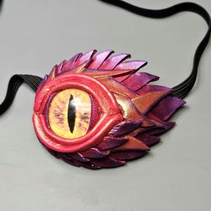 May include: A fantasy-themed eye patch with a dragon eye design. The eye patch features a vibrant, multi-colored design with shades of red, purple, and gold. The eye itself is yellow with a black slit pupil. A black elastic band is attached.