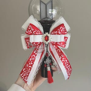 May include: A black and clear light stick with the letters "BTS" on top. The light stick is decorated with a red and white bow, a red jewel, and a red tassel. The bow has a decorative pattern.