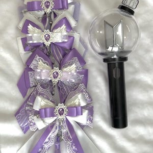May include: A collection of purple and white decorative bows with jeweled accents and a black and clear light stick. The bows feature lace and ribbon details. The light stick has a black handle and a clear orb with a silver logo.