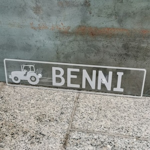 Può includere: Un cartello in acrilico trasparente con una silhouette di trattore bianca e il nome "BENNI" in lettere bianche.