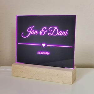 Könnte beinhalten: Ein schwarzes Acrylschild mit einem Holzsockel. Das Schild hat eine pinkfarbene Neon-Inschrift mit dem Text "Jan & Dani" mit einem Herzsymbol und dem Datum "08.06.2024".